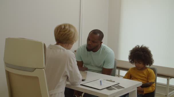Médico profesional que explica el tratamiento médico al padre negro preocupado durante la visita al hospital — Vídeo de stock