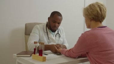 Afrikalı erkek pratisyen doktor ofisteki orta yaşlı kadının nabzını kontrol ediyor.