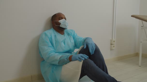 Fáradt afrikai férfi sebész ppe ül a padlón, pihenés után hosszú műtét a kórházban — Stock videók