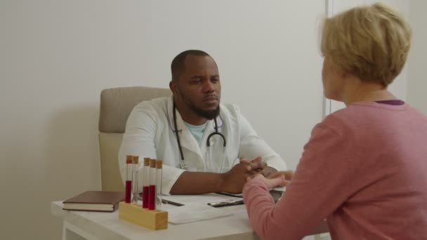 Зріла жінка-пацієнтка скаржиться на стан здоров'я до уважного чорного лікаря в клініці — стокове відео