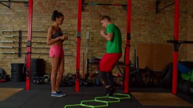 Spor hocası, çeviklik merdiveni kullanarak siyah formda bir kadına kardiyo çalışması gösteriyor.