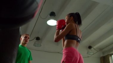 Spor formundaki siyah bayan boksör antrenörle içeride kum torbası yumrukluyor.