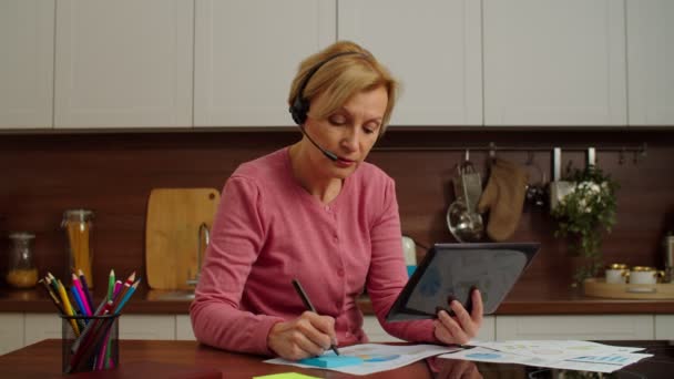 Pretty mature businesswoman in headset with tablet pc working remotely from home — Wideo stockowe