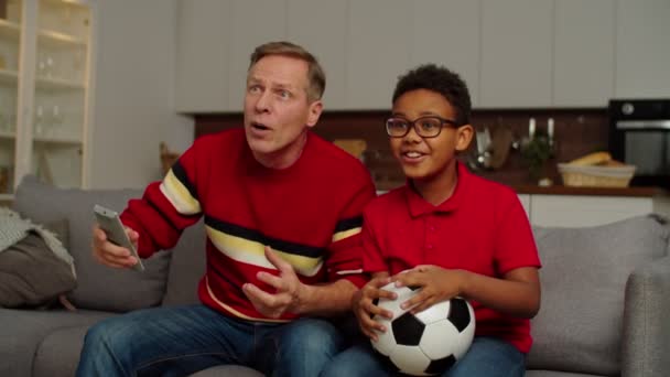 Opgewonden grootvader en leuke school leeftijd multiraciale jongen kijken voetbal op tv — Stockvideo