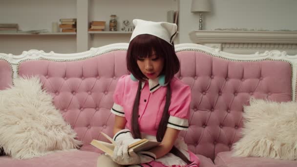 Belle asiatique femme en femme de ménage cosplay costume lecture un livre sur canapé — Video