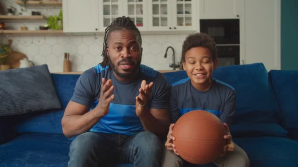 Glad afroamerikansk far och son tittar på basket match på tv hemma — Stockvideo