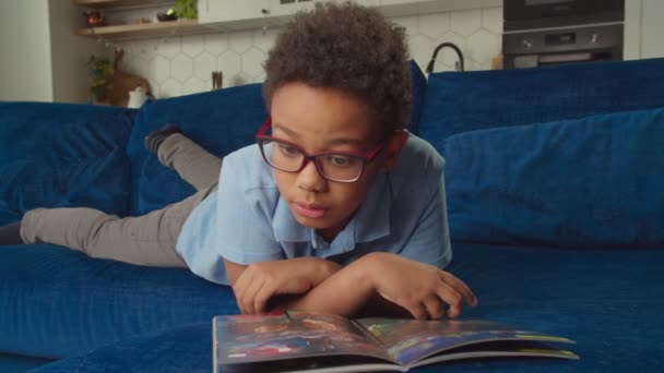 Cute preadolescent African American boy in eyeglasses reading comics on sofa — Stock Video