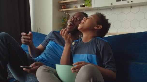 Glad tonåring svart pojke och pappa koppla av på soffan och titta på film på tv — Stockvideo
