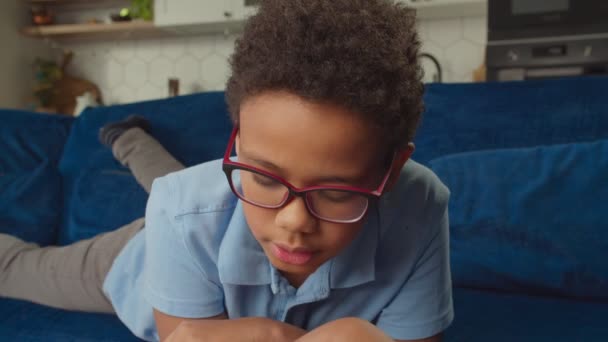 Έξυπνος εστιασμένος Αφρικανός μαθητής με γυαλιά που διαβάζει στον καναπέ. — Αρχείο Βίντεο