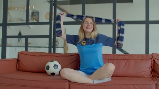 興奮した魅力的な中年女性サッカーサポーターでチームジャージーでスカーフを見ながらサッカーゲームでテレビ 応援チームとチャットスローガンながら 国内の部屋でソファに座って — ストック動画