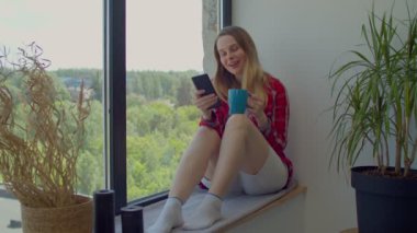 Kaygısız çekici yetişkin kadın pencere kenarında oturuyor, kahveyi yudumluyor ve internete bağlanıyor, sosyal medya içeriğini araştırıyor, cep telefonu kullanıyor ve iç mekanlarda eğleniyor..