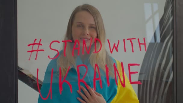 一位积极可爱的中年妇女 裹着乌克兰国旗 望着窗外 站在玻璃上写着乌克兰 呼吁人们在战争期间支持乌克兰 并展示自己的标志 — 图库视频影像