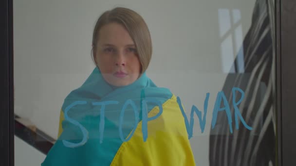 悲しい魅力的な中年の女性は ウクライナの平和を望むウクライナのフラグに包まれました 内部的にガラスに書かれたテキストを停止戦争でウィンドウを介して動揺式を探しています — ストック動画