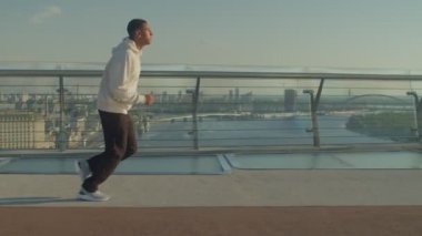 Motive olmuş yakışıklı sporcu kapüşonlu Afrikalı erkek koşucunun yan görünüşü kardiyo antrenmanı yapıyor, sabah Ray 'le yaya köprüsünde koşuyor ya da şehrin arka planında sıcak gün doğumu yapıyor.