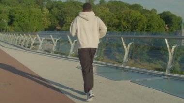 Spor kıyafetleri ve kapüşonlu bir sporcunun yaya köprüsünde koşup, yükselen sıcak güneş ışınları altında kardiyo antrenmanı yapması..