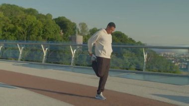 Kararlı spor sporunun portresi. Siyah adam bacak egzersizleri yapıyor, ısınıyor, kaslarını kasıyor ve kaslarını kasıyor. Sabah gündoğumunda şehir sokaklarında koşmadan önce..