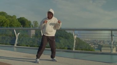 Yoğunlaşmış motivasyonlu spor kaslı siyah kapüşonlu erkek boksör vücut egzersizi için ısınma egzersizi yapıyor, şafak vakti yaya köprüsünde gölge boksu yaparken yumruk tekniklerini geliştiriyor..