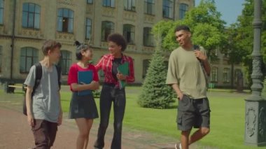 Positive attractive diverse multiethnic college friends with backpacks saying goodbye to each other, embracing and fist bumping at university campus after finishing lesson.