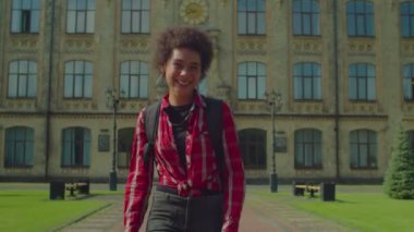 Sırt çantasıyla kampüste yürüyen, neşeli gülümseyen, neşeli ruh hali ve mutluluğu ifade eden pozitif Afrikalı kız üniversite öğrencisi..