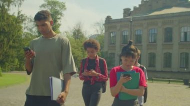 Portrait of attractive diverse multiracial college friends phubbing each other, networking online on smartphones while walking together to study at university campus.