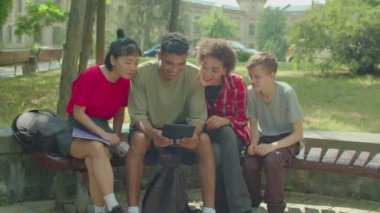 Positive beautiful diverse multicultural young people sharing digital tablet, networking and browsing social media content online while enjoying leisure, sitting on bench in public park.