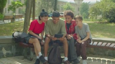 Joyful diverse multiethnic college friends checking online exam results on tablet pc, celebrating academic success with fist pump gestures, expressing excitement and happiness while sitting on bench.