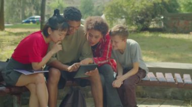 Attractive diverse multiracial college students checking exam results online using digital tablet, expressing positive and negative emotions while sitting on bench outside university building.