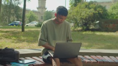 Thoughtful stylish handsome African male student learning, networking online on academic project using laptop pc while sitting on bench at university campus.