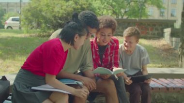 Portrait of smart beautiful diverse multiethnic college students with textbooks learning together, preparing for university exam while sitting on bench at campus.