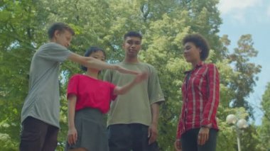 Positive attractive diverse multiethnic young people standing in circle, stacking hands together, showing friendship, unity and teamwork on sunny day in public park. Low angle view.