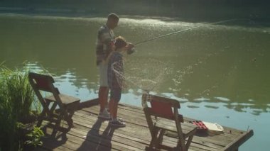 Positive handsome African dad and excited school age stepson enjoying recreational activity and fishing by pond , catching fish with spin fishing rod while relaxing together at summertime at sunrise