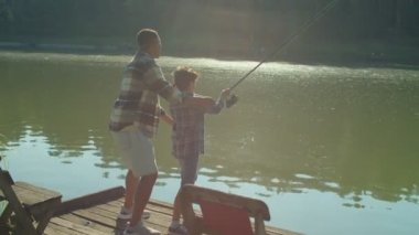 Caring handsome African American dad teaching cute preadolescent son to fish and cast spin fishing rod, explaining fishing techniques while multiethnic family enjoying leisure on pond at dawn.