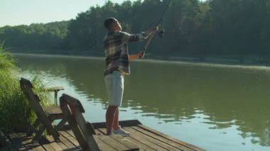 Skillful handsome black male fisherman standing on wooden jetty, casting spin fishing rod while enjoying leisure and fishing on lake in rays of warm summer sunrise.