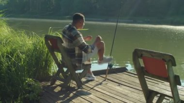 Rear view of excited attractive African male fisherman with spin fishing rod , catching freshwater fish while enjoying fishing on wooden pier on pond in rays of rising sun.