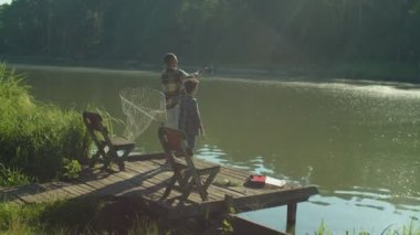 Cheerful handsome African American man and adorable preadolescent Middle Eastern stepson standing on wooden pier, casting fishing rod while family enjoying fishing by lake at sunrise.