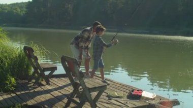 Joyful cute school age boy, with help of caring handsome African American stepfather, catching fish with spin fishing rod while family enjoying leisure and fishing on wooden pier on pond at sunrise.