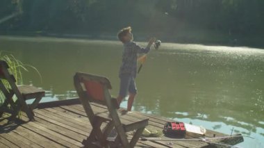 Positive skillful adorable preadolescent Middle Eastern boy standing on wooden pier, casting spin fishing rod while enjoying leisure and freshwater fishing on lake at daybreak
