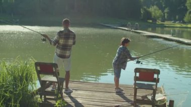 Rear view of happy preadolescent Arab boy and attractive African stepfather standing on wooden jetty, casting fishing rods while united multicultural family spending leisure fishing on pond.