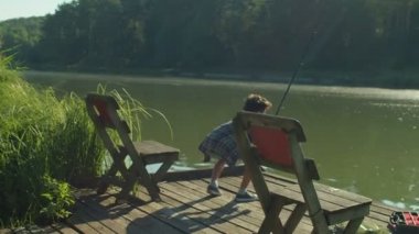 Cute school age Arab boy catching freshwater fish with spin fishing rod on wooden jetty on pond, expressing disappointment and irritation about fish got away while fishing in morning.