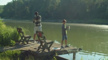 Positive attractive African American father and adorable preadolescent son standing on wooden jetty, casting spin fishing rods while multiracial family enjoying fishing on pond at sunrise.