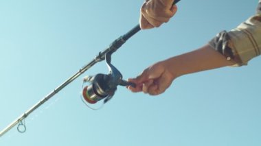Low angle view of black male hands holding rod and turning fishing reel over blue sky background during angling on summer day. Close up.