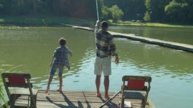 Rear view of happy cute preadolescent boy and attractive African father standing on wooden jetty, casting fishing rods while united multicultural family spending leisure angling on pond.