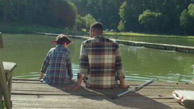 Rear view of positive African American father and cute multiracial teenage son sitting on wooden pier on pond , friendly talking and sharing, expressing strong relationship and bond during fishing.