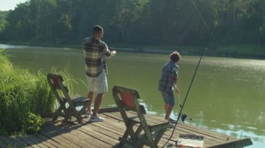 Positive African American father with spin fishing rod standing on wooden jetty, catching fish while excited cute Middle Eastern teenage stepson helping with landing net during fishing on pond.