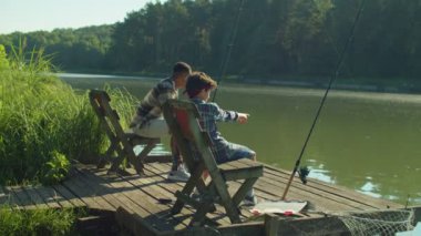 Handsome African stepfather and cute school age Middle Eastern boy sitting on wooden pier, friendly talking and bonding while multiethnic family enjoying fishing on lake at dawn.