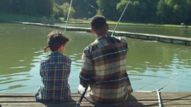 Rear view of multiracial teenage son and African dad spending weekend activity, talking and fishing with fishing rods from wooden jetty on lake while multicultural family enjoying leisure in nature.