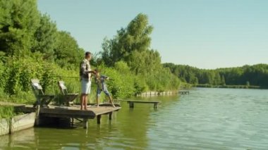 Happy handsome black father and cute preadolescent multiethnic boy standing on wooden pier, casting spin fishing rods and angling on pond while multiracial family enjoying leisure in summertime.