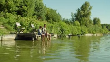 Cheerful handsome African American father and happy lovely preadolescent multicultural teenage son sitting with fishing rods on wooden pier, dangling feet into water and angling on summer day.