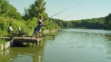 Joyful adorable school age multiracial boy and positive black father casting fishing rods from wooden pier on pond, talking and enjoying fishing together on summer day.