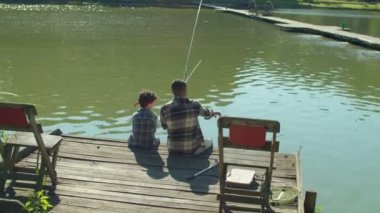 Positive African American father and cute preadolescent multiethnic son with fishing rods sitting on wooden pier, chatting and angling on pond while multiracial family enjoying leisure on summer day.